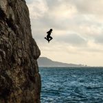 Side view of unrecognizable traveler jumping into wavy ocean from massive rocky cliff against cloudy sunset sky